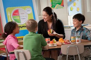 photo of teacher with three first graders sitting around a small table of a variety of food items