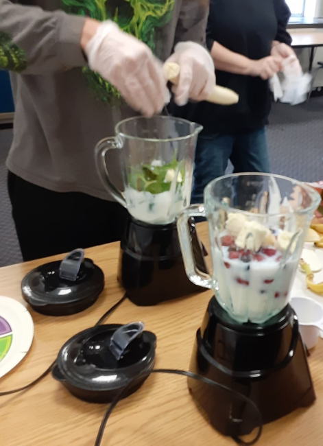 close up image of two blenders and two youth's hands putting slices of bananas into the blenders that contains a variety of fruits, vegetables, milk and yogurt.