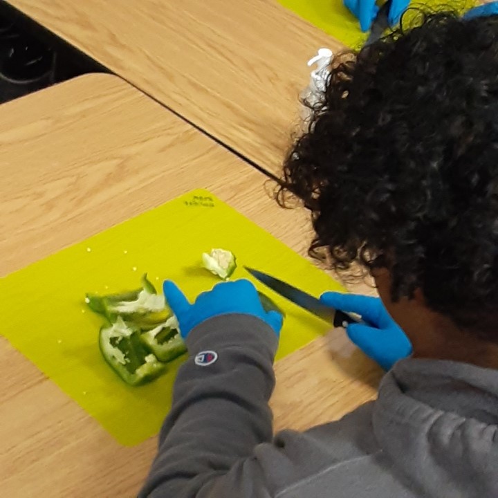 image looking over the shoulder of a teen youth slicing green peppers on a yellow cutting mat