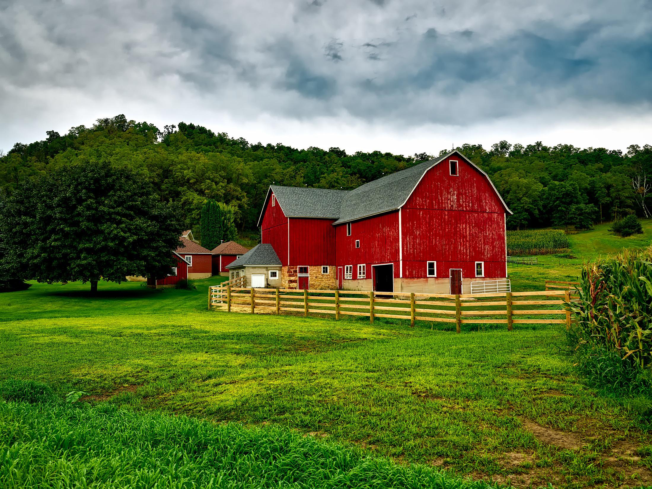 image of a red painted barn with dark gray shingled roof, positioned against a tree background with out buildings, small corn patch and garden area. the barn has a stone and cement base and has a natural wood fence that extends from one barn corner out and around a small pasture area, reconnecting to the opposite corner of the barn