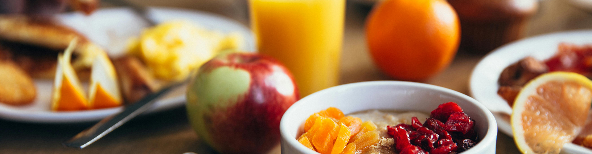 close up of healthy breakfast options with a cup of fruit, an apple, an orange, a plate of sliced fruit and a glass of orange juice