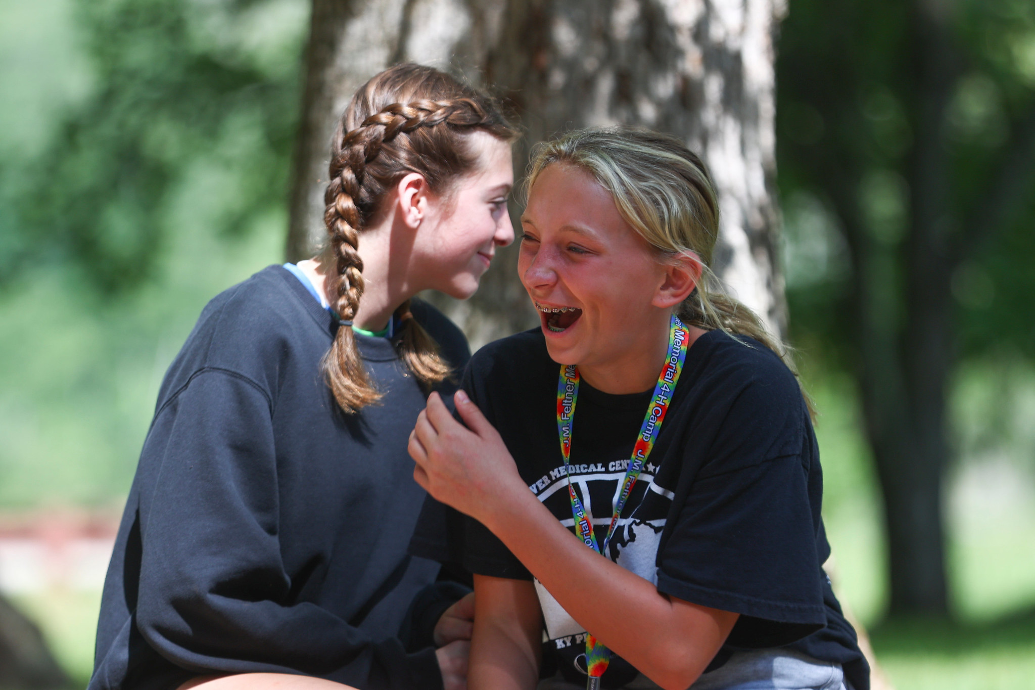 two young teen girls sharing a laugh in the shade of a nearby tree